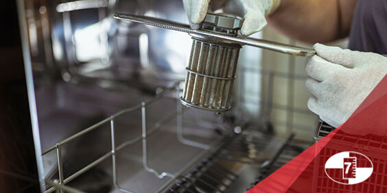 Person wearing protective gloves removing a cylindrical filter from the bottom of an open dishwasher.