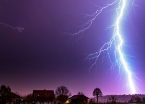 A lightning bolt strikes above a house, illuminating the night sky with dramatic flashes.