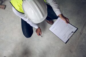 A man wearing a hard hat and safety vest holds a clipboard, standing in a construction environment.