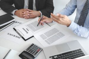 Two business professionals seated at a table, reviewing papers and working on a laptop together