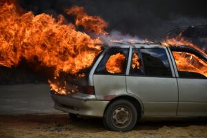  A tan sedan with broken out windows is on fire, with flames protruding from the interior of the vehicle.