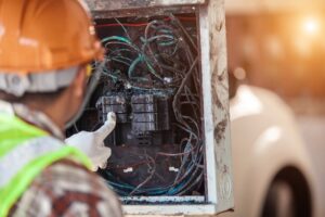 A person in a hard hat and safety vest inspecting fire-damaged electrical wiring and components inside an electrical panel